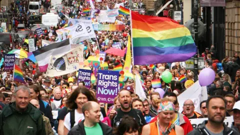 People in the Edinburgh Pride march walk along a road holding rainbow flags and trans rights signs 