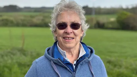 Kate has curly grey hair and round sunglasses. She wears a blue hooded top and stands in front of the fields where the solar farm will go. 