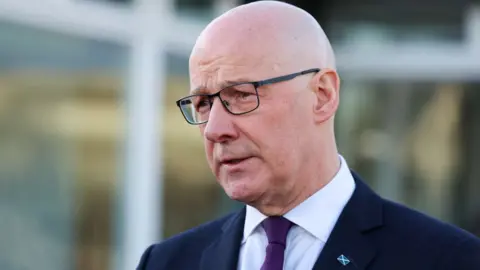 John Swinney, who is bald with glasses, looks to his right in a close-up shot. He is wearing a dark suit and white shirt with a purple tie. He is standing outside, with windows out of focus behind him. 