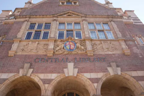 City of Wolverhampton Council The main entrance of Wolverhampton Central Library seen from street level, looking up at the ornate first-floor façade. A colourful royal coat of arms sits centrally above the 'Central Library' lettering, flanked by carved stone lions. 