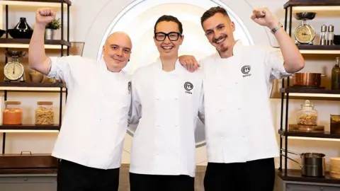 Three people wearing chef whites and smiling at the camera. A woman with dark hair and glasses is flanked by a bald man and a man with short dark hair and a beard who are both pumping their fists.