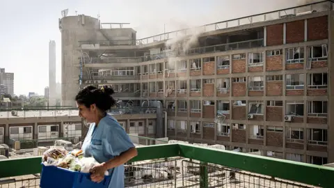 Image shows a nurse carrying medical supplies past a building with smoke billowing out at Soroka Hospital following an Iranian missile attack in Beersheba, southern Israel, on 19 June, 2025
