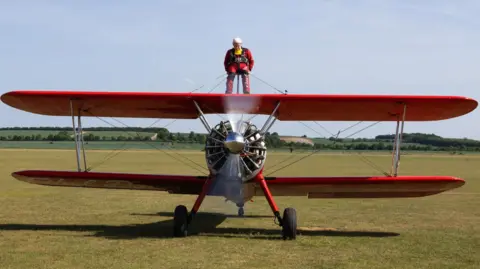Guinness World Records Harry Heasman, in a red and black jump suit, is shown atop a red biplane. The plane's singular propeller engine is shown in motion on a grass landing strip at the airfield.