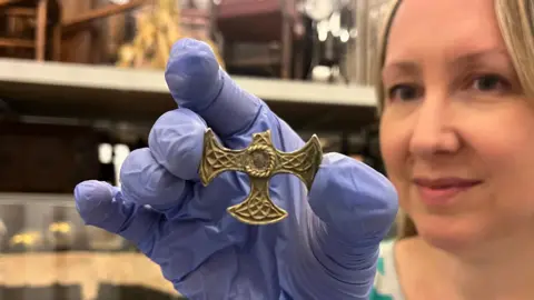 A woman holds a small gold cross with the top arm missing. She is wearing blue gloves. She has blonde hair. The cross is about 5cm wide