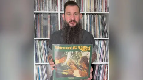 Martin is holding Billy Nicholls record, would you believe.  The cover is green with yellow text.  The image is a wavy effect photo of the main artists involved. Martin has dark hair and a very long beard. He is sat infant of loads of vinyl records in shelves 