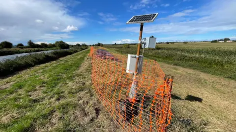 Monitoring equipment on the riverbank at Fiskerton. A pole has a solar panel at the top used to the power the equipment below. 