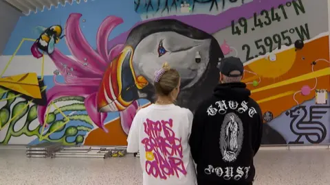 Tiffany Anna and Woodie are looking at he mural with their backs to the camera.