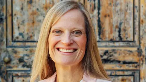 Worcester Theatres A woman with long blonde hair smiles widely as she stands in front of an old brown door.