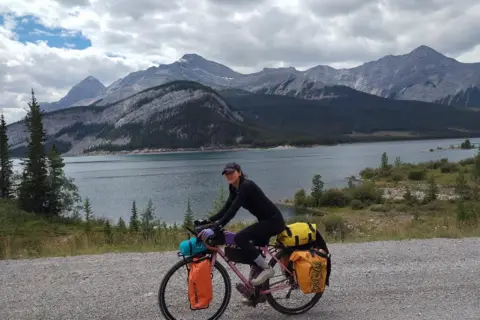 Naomi Hancocks Naomi Hancocks is sitting on her pink bike with scenic mountains and a lake in the background. She has many panniers on her bike.