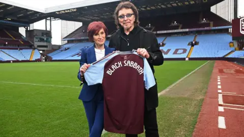 Getty Images Sharon Osbourne and Tony Iommi are standing on the green pitch of Villa Park, with stands and blue seats in the background. They are holding up an Aston Villa football shirt that says "Black Sabbath" on the back.