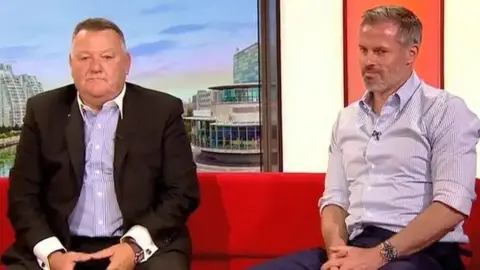 Mark King is sat alongside Jamie Carragher on a red sofa in the BBC Breakfast TV studio. Mark King is wearing a lilac and white striped shirt and a black suit jacket. Jamie Carragher is wearing a lilac and white striped shirt with the sleeves rolled up.