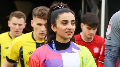 Sabriah Nawroozi walks out of the tunnel at a football stadium with football players from Harrogate Town AFC.