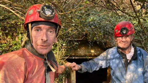 Tim Clarke in a red waterproof suit looking at the camera beside a mine opening, and Pat Moret in a blue suit. Both men are wearing red hard hats with torches on the front of them