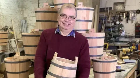 Alan Paulus Alan Paulus wearing a red jumper and glasses. He is holding a bucket and is stood in front of a pile of buckets in his workshop.