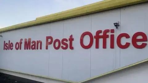 Isle of Man Post Office in large red lettering on the side of the roof of a post office building the surface is white with yellow painted guttering.