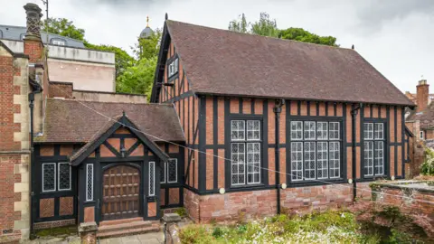 Historic England The exterior of a red brick building with black wooden beams, large windows and a brown tiled roof. There is a covered entrance with a large wooden door on the left hand side