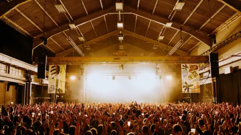 Prospect A large warehouse room filled with a big crowd of people. They are facing a stage that is lit up by very bright yellow stage lights.