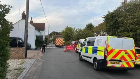 Tony Fisher/BBC A residential street is partially blocked by emergency vehicles and barriers. A white van marked “Scientific Services Unit” is parked on the right side of the road, alongside other response vehicles with high-visibility markings. Red-and-white tape and several traffic cones are set up around a “Road Closed” sign, preventing access further down the street. A uniformed officer stands near the cordon. In the background, additional vehicles and personnel are visible near houses and trees, with overhead utility wires crossing the sky.