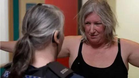 Adriana Orme, who is wearing a black sports bra, holds both arms out to the side, as she is arrested by a female police officer. The officer is wearing black police uniform and has her hair in a ponytail.