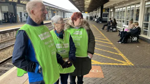 Three Samaritan volunteers stood to the left of the platform. There are passengers sat on benches to the right. The volunteers are wearing green bibs with the Samaritans branding and number below. 