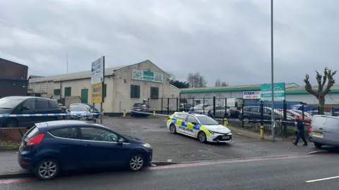 BBC A police car and several other vehicles parked outside a white building with green metal grilles over the windows and a white sign painted in green with indistinguishable English and Arabic text. A sign on the pavement reads Jamia Masjid, along with Arabic text.