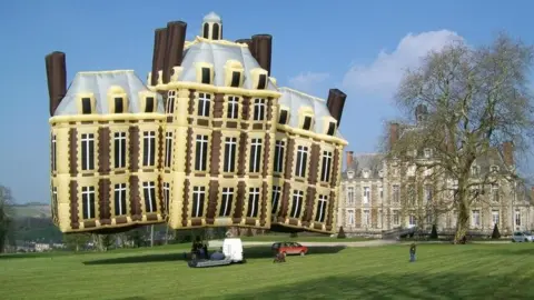 Longleat A large hot air balloon in the shape of a brown, sand and white chateau, commissioned by Malcolm Forbes, publisher of Forbes magazine. It is tethered on a green lawn with a large country house in the background.