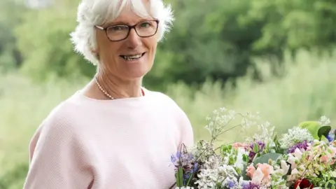 Andrea Gilpin A bespectacled Gill Hodgson wearing a pink top and holding a large bunch of flowers standing in an outdoor location with trees in the background.