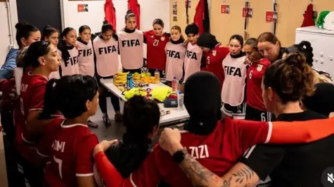 Afghan Women United About 20 women huddle in a circle in a changing room with their arms around each other. Some wear red football shirts and some have white bibs over their kit with the word Fifa written in dark blue. There is a table in the middle of the circle with a few bottles and some fruit on it.