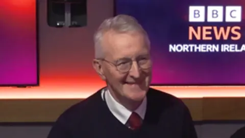 BBC Hilary Benn sitting in Radio Ulster studio booth in front of a microphone. He is wearing a dark pullover, white shirt and red tie. He has round glasses and short grey hair. Behind him are two screens with the BBC News Northern Ireland logo on them on purple/red backgrounds.