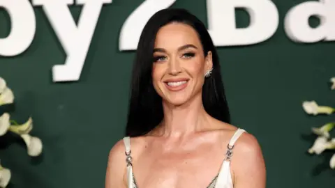 Close-up shot of pop star Katy Perry in a white dress, wearing her long black hair down, against a dark green background
