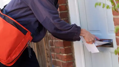 Getty Images Close up of a man wearing a navy postman's jacket with a red bag slung over his shoulder pushing several white envelopes through the letterbox on a white door. The picture is cropped to only show the postman's arms and torso. This is a stock photo.