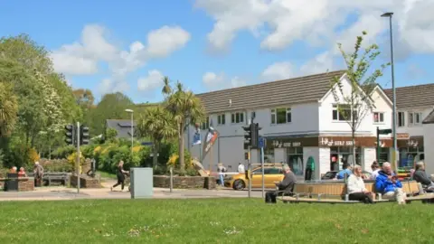 Artist's impression of a grass area with shops and open air seating behind and people sitting on benches. Traffic lights and part of a road are visible as well as trees and distant countryside are also visible. 