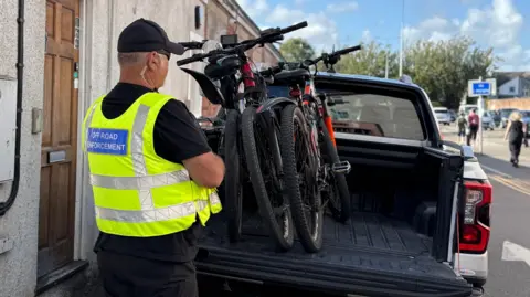 Three mountain bikes loaded into the back of a pick-up truck. Next to the vehicle a council officer stands with his back to the camera, wearing a black T-shirt and yellow hi-vis jacket which has a blue label on the back which says "Off Road Enforcement" 