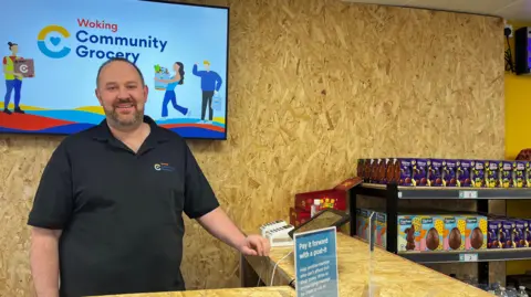A man wearing a black shirt stood behind a desk. A TV in the background has a graphic reading "Woking Community Grocery".