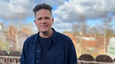 Jon Wright/BBC Luke Wright standing outside with a backdrop of trees and clouds.