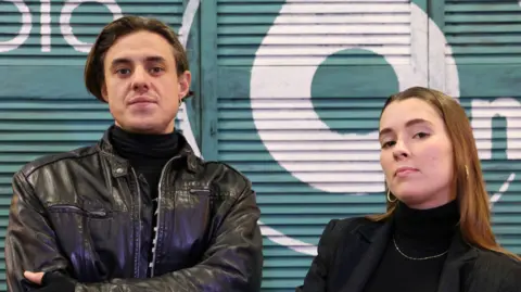 Aidan Moore and Grace Stephenson AKA Sugar Bones and Janet Planet of Australian band Confidence Man - they are both looking directly into the camera with their arms folded.
He has short dark hair and is wearing a leather jacket, she has shoulder length brunette hair and a dark jacket and top on.