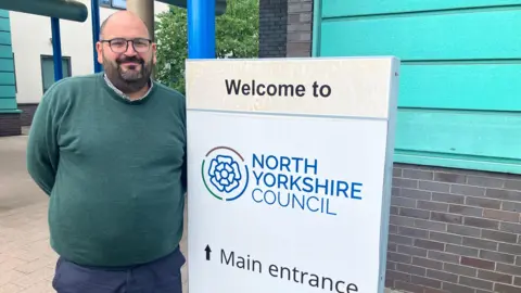 BBC/Richard Edwards A man in a green jumper and blue trousers stands outside North Yorkshire Council's offices in Selby. He is wearing glasses, smiling at the camera and has a black beard.
