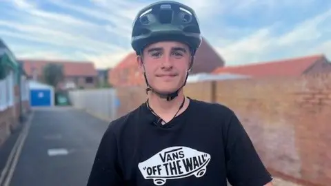 A teenage boy is wearing a black top with a white skateboard logo which reads: 'VANS OFF THE WALL'. He is wearing a dark green helmet and smiling into the camera. You can see a path behind him leading to what looks like a garage and on the right is a red brick wall.
