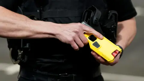 A close up of a police officer holding a taser in his hands. The taser is yellow and black with two red pins.  