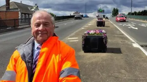 A man with grey hair wears a suit under an orange hi-vis jacket and stands in front of a planter in the central reservation on a dual carriageway. There are several black planters behind him featuring colourful artificial flowers. 