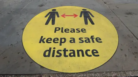 Covid-era social distancing warning sign on a pavement. It is a circular yellow sign with the words written in black: "Please keep a safe distance".