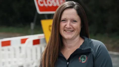 John Fairhall/BBC Kim Turner is wearing a dark zip-up jacket. She has long brown hair and is smiling while standing in front of various road signs and barriers.