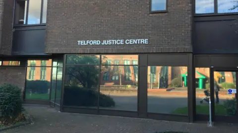 BBC A nondescript looking court building. It is brick and brown and we can see reflective windows and an automatic door. A woman with a pushchair can be seen in the image. The words Telford Justice Centre are on the front of the building. 