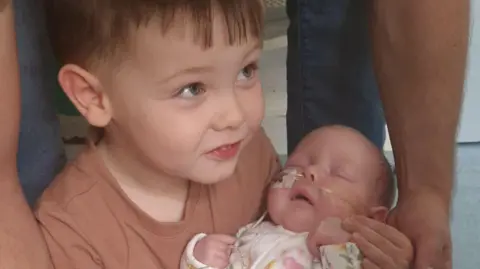 Maxwell is holding his baby sister Lily- who has tubes in her nose. 