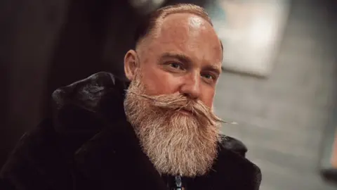 A man with a bushy beard and a waxed pointy moustache is looking at the camera. He is wearing a black top