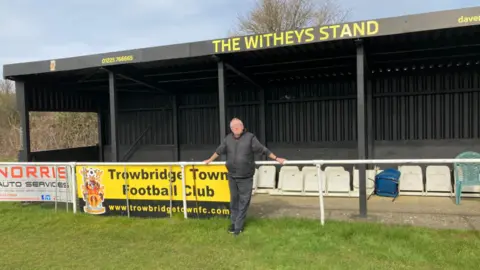 Trowbridge Town FC A man wearing black clothes stands in front of a series of plastic seats in a football club. He is under a sign in which 