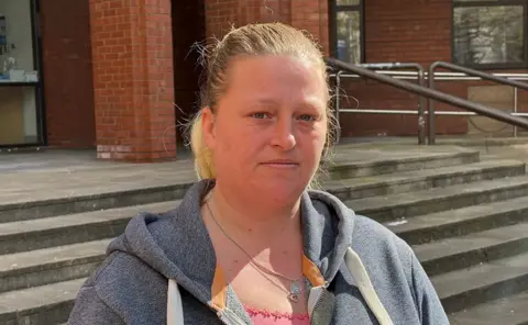 Michaela Baldwin stands outside Hull Crown Court. She is in front of a brick building with concrete steps. She has light hair pulled back into a ponytail and is wearing a grey hoodie over a pink top, with two necklaces visible. She looks directly at the camera with a neutral expression in bright daylight.