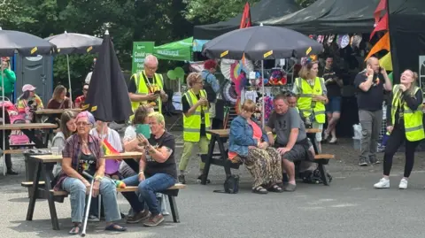 People are sitting on wooden benches and some people are walking about with yellow high-vis jackets. There are stalls in the background selling items.