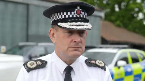 Jamie Niblock/BBC Ben-Julian Harrington is wearing a white shirt, black tie, black hat and has black epaulettes on his shoulders. A marked police car is parked behind him.