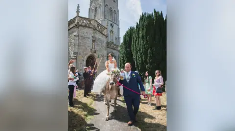 Bride in white wedding dress being led out of a church while riding a pale-coloured cow. The cow has a flower garland on its head and is being led by a man holding a pink leash. A guard of honour can be seen behind comprised of people in suits raising farming tools into the air.